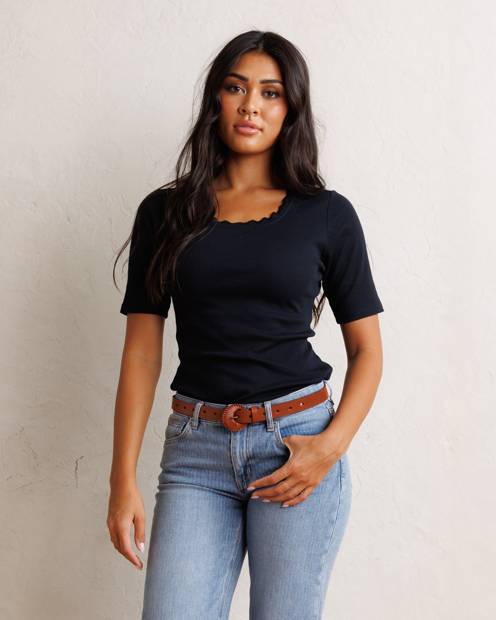 A woman modeling a short-sleeve black top with a scalloped neckline, paired with light blue jeans and a brown belt, standing against a textured off-white wall.