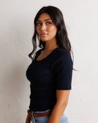 A woman with long dark hair wearing a black short-sleeve scoop-neck t-shirt and light blue jeans with a brown belt, standing against a textured off-white wall.