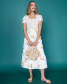 Woman wearing a white floral dress holding a woven bag against a teal background