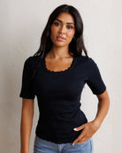 A woman modeling a black short-sleeve crewneck t-shirt with a scalloped scoop neckline, paired with blue jeans, standing against a textured off-white wall.