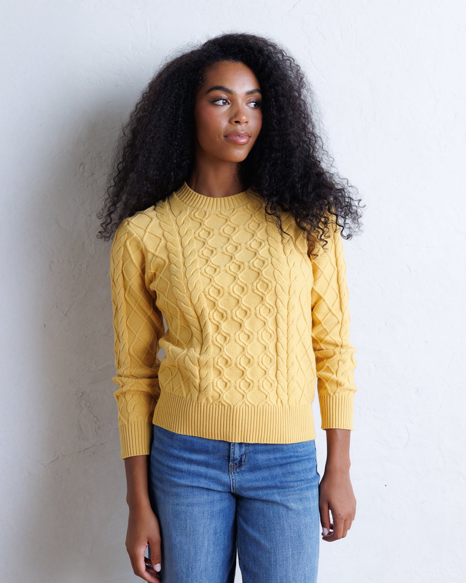 A woman with long, curly black hair wearing a yellow cable-knit sweater and blue jeans, standing against a white textured wall.