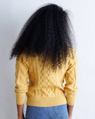 A person shown from the back wearing a mustard yellow cable-knit sweater and blue jeans, with very large dark curly hair, against a plain white wall.