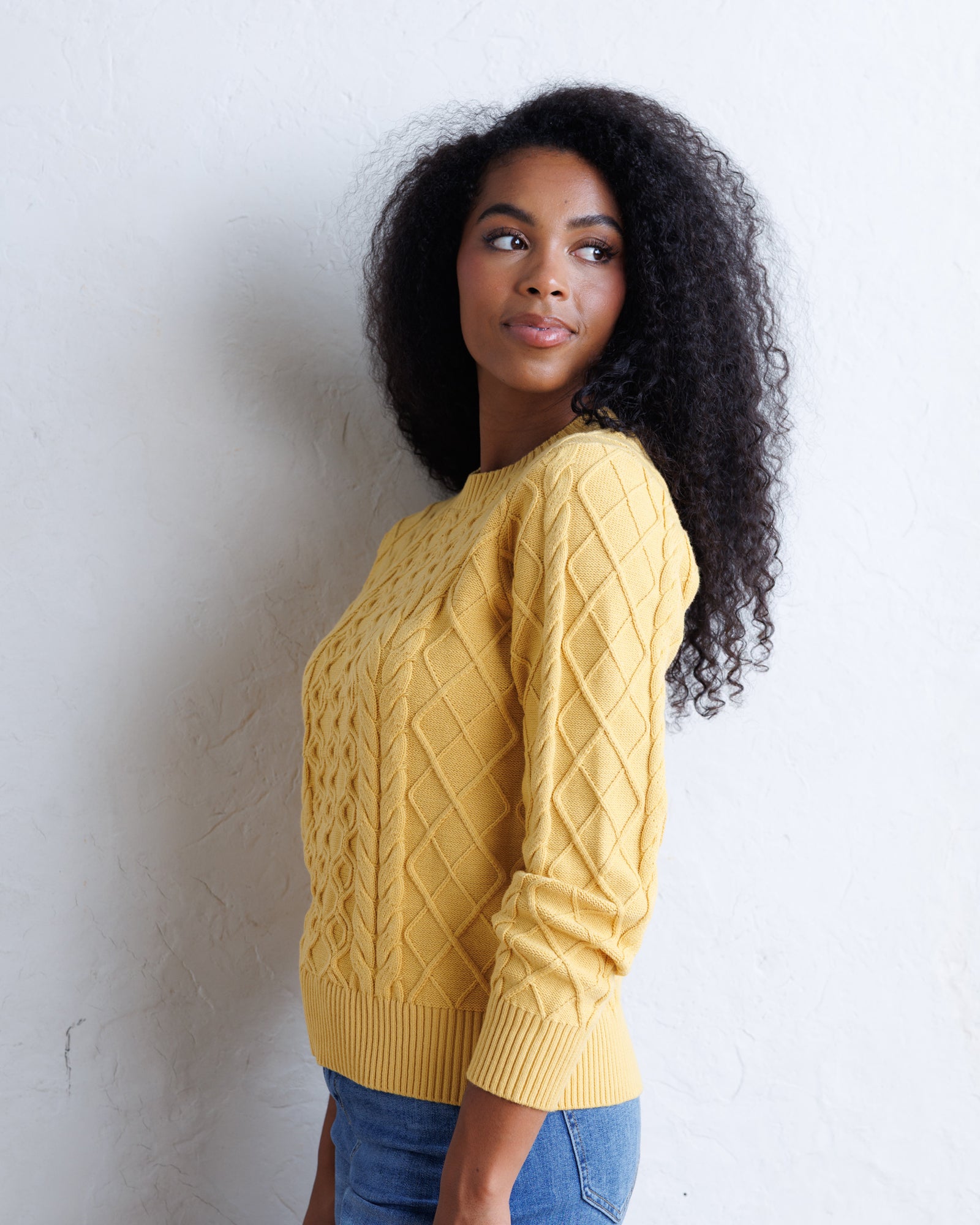 A woman with curly black hair wearing a mustard yellow cable-knit sweater and blue jeans, standing against a white textured wall.