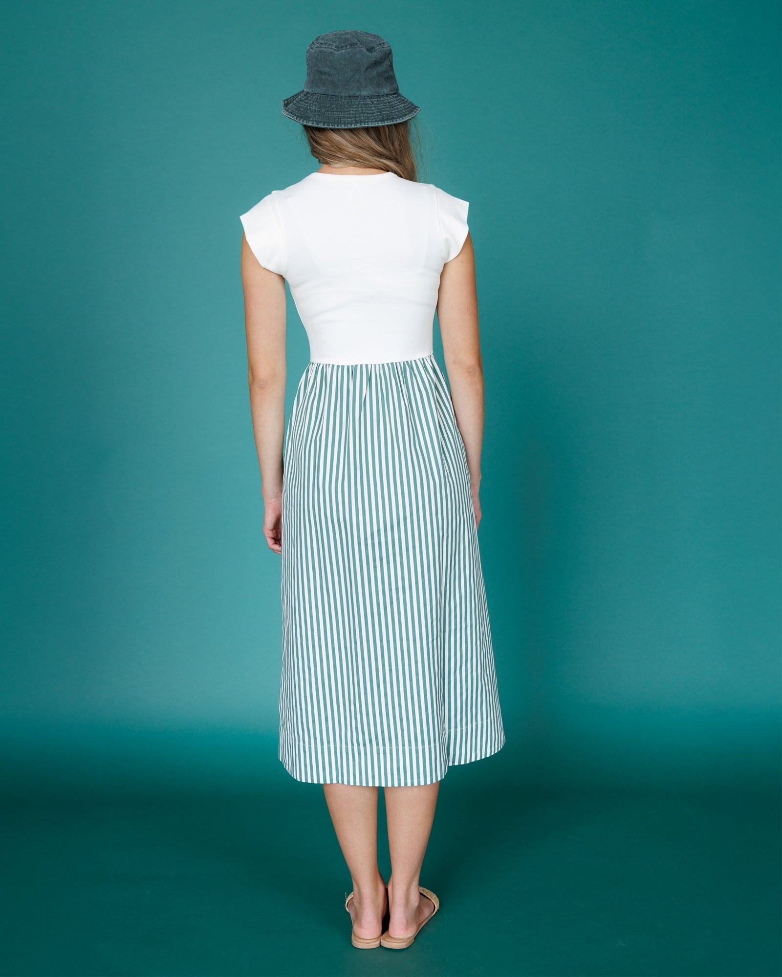 Woman wearing a white top and striped skirt with a blue hat on a teal background