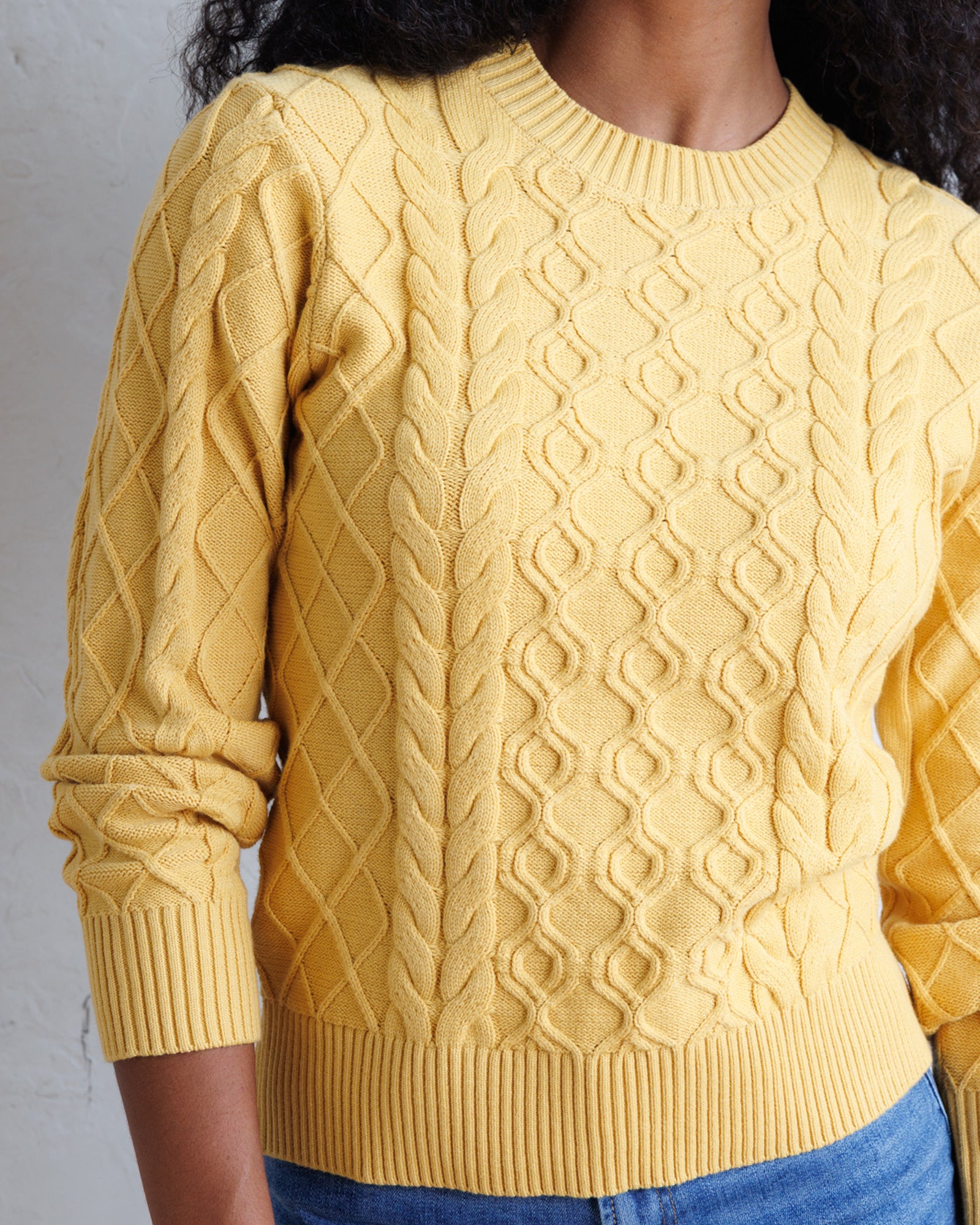 A close-up of a person wearing a textured yellow cable-knit sweater with a crew neck and ribbed hem, paired with blue jeans against a light gray wall.