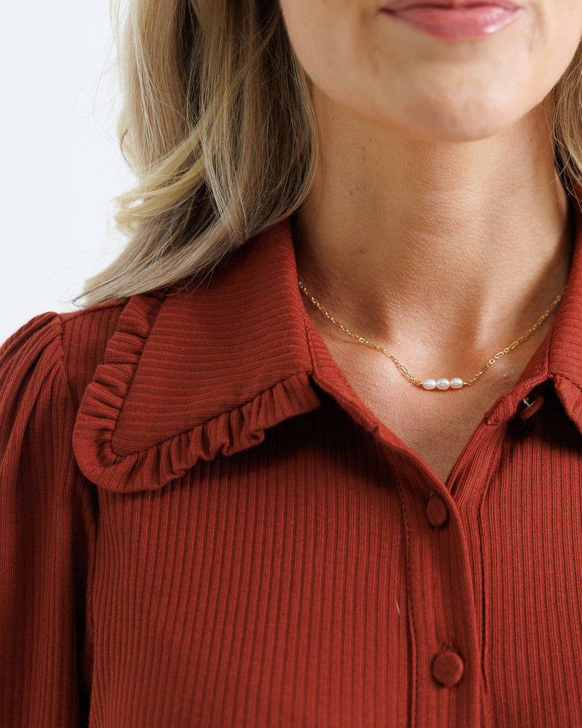 Close-up of a person wearing a rust-colored blouse with a ruffled collar and a gold necklace with pearls.