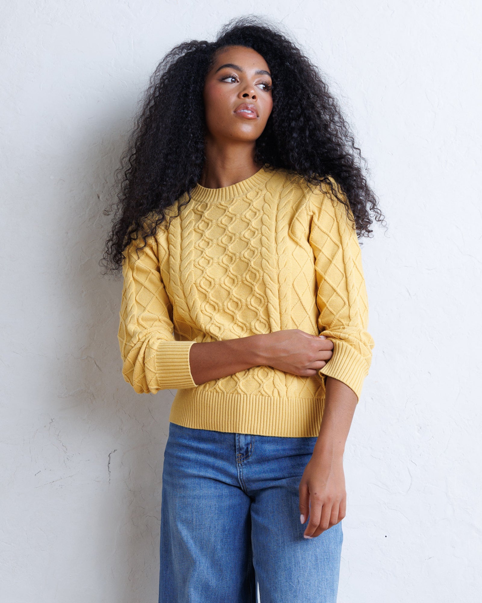 A woman with long, curly black hair wearing a yellow cable-knit sweater and blue jeans, standing against a white textured wall.