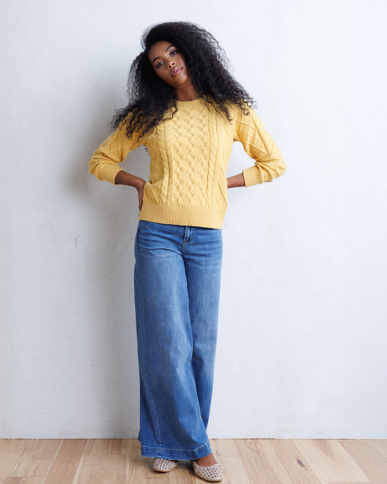 A woman with long, curly black hair stands against a white wall wearing a yellow cable-knit sweater and wide-leg blue jeans.