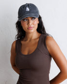 A woman wearing a dark gray baseball cap with a small white ghost graphic and a brown ribbed tank top, posing against a white textured wall.
