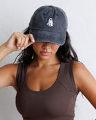 A woman wearing a dark gray washed baseball cap with a small white embroidered ghost on the front, holding the brim with one hand while wearing a brown ribbed tank top against a light gray wall.
