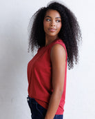 A woman with long, curly black hair is shown from the side, wearing a sleeveless red top and dark blue jeans against a white textured wall.
