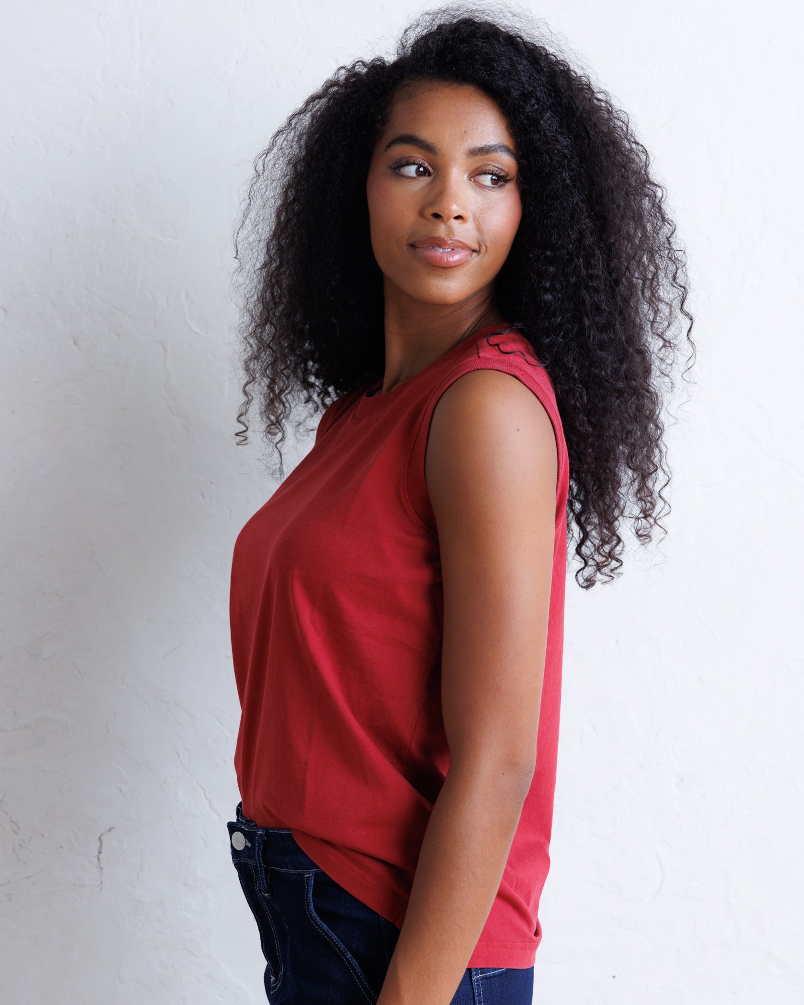 A woman with long, curly black hair is shown from the side, wearing a sleeveless red top and dark blue jeans against a white textured wall.
