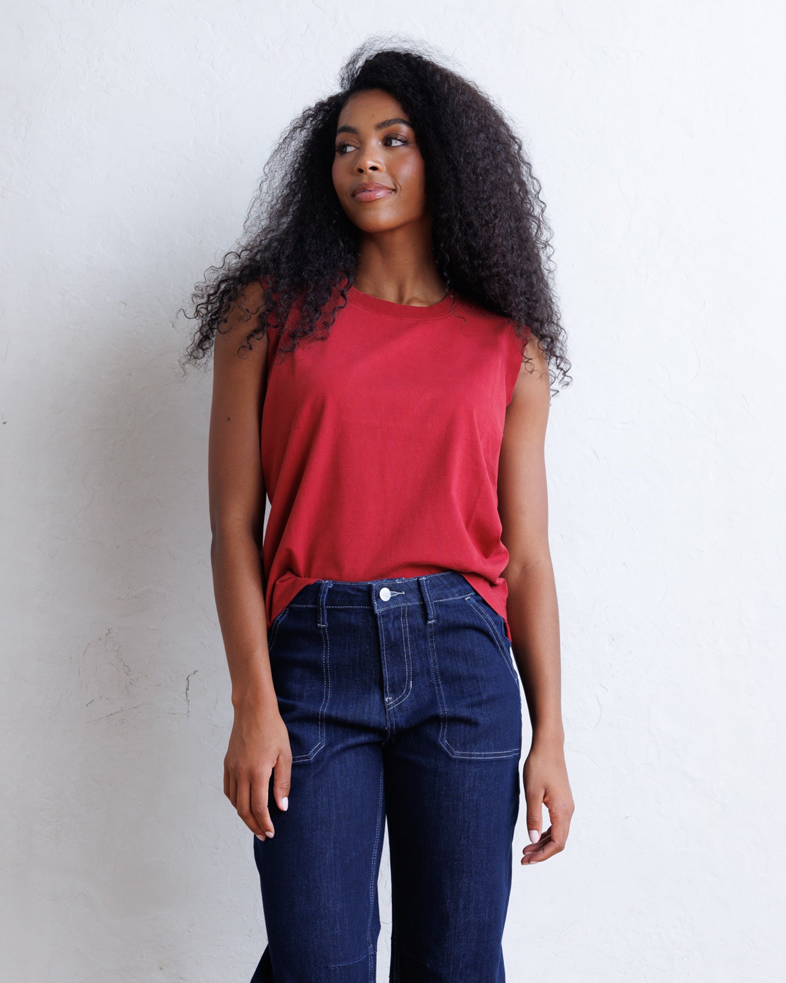 A woman with long, curly black hair wearing a sleeveless red top and dark blue jeans, standing against a textured white wall.