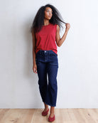 A woman with curly black hair stands against a white wall wearing a sleeveless red top, dark blue cropped jeans, and red flats.