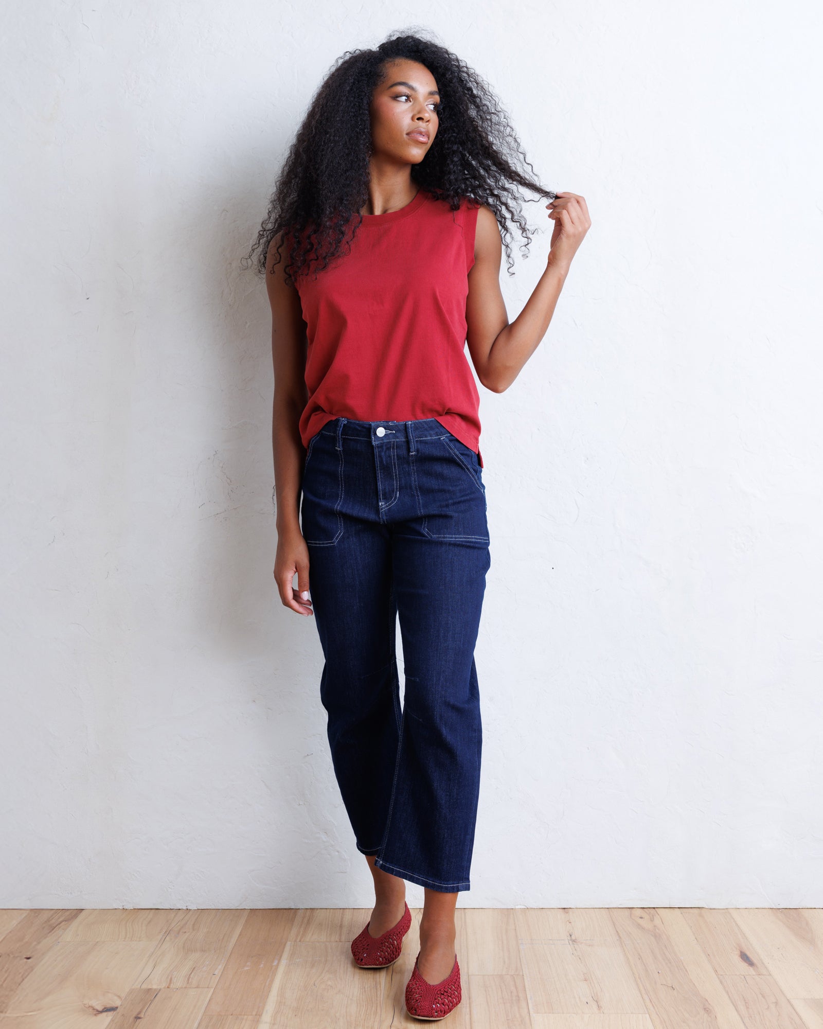 A woman with curly black hair stands against a white wall wearing a sleeveless red top, dark blue cropped jeans, and red flats.