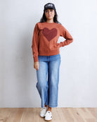 A woman stands against a light gray wall wearing a rust-orange crewneck sweatshirt with a large dark red heart on the front, blue jeans, white sneakers, and a black baseball cap with 'CIRA' in white text.