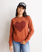 A woman wearing a rust-orange long-sleeve sweatshirt with a dark red heart patch on the chest, blue jeans, and a black baseball cap, posing against a textured off-white wall.