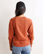 A woman viewed from behind wearing a rust-orange raglan crewneck sweatshirt, a black trucker hat, and blue jeans against a plain white wall.