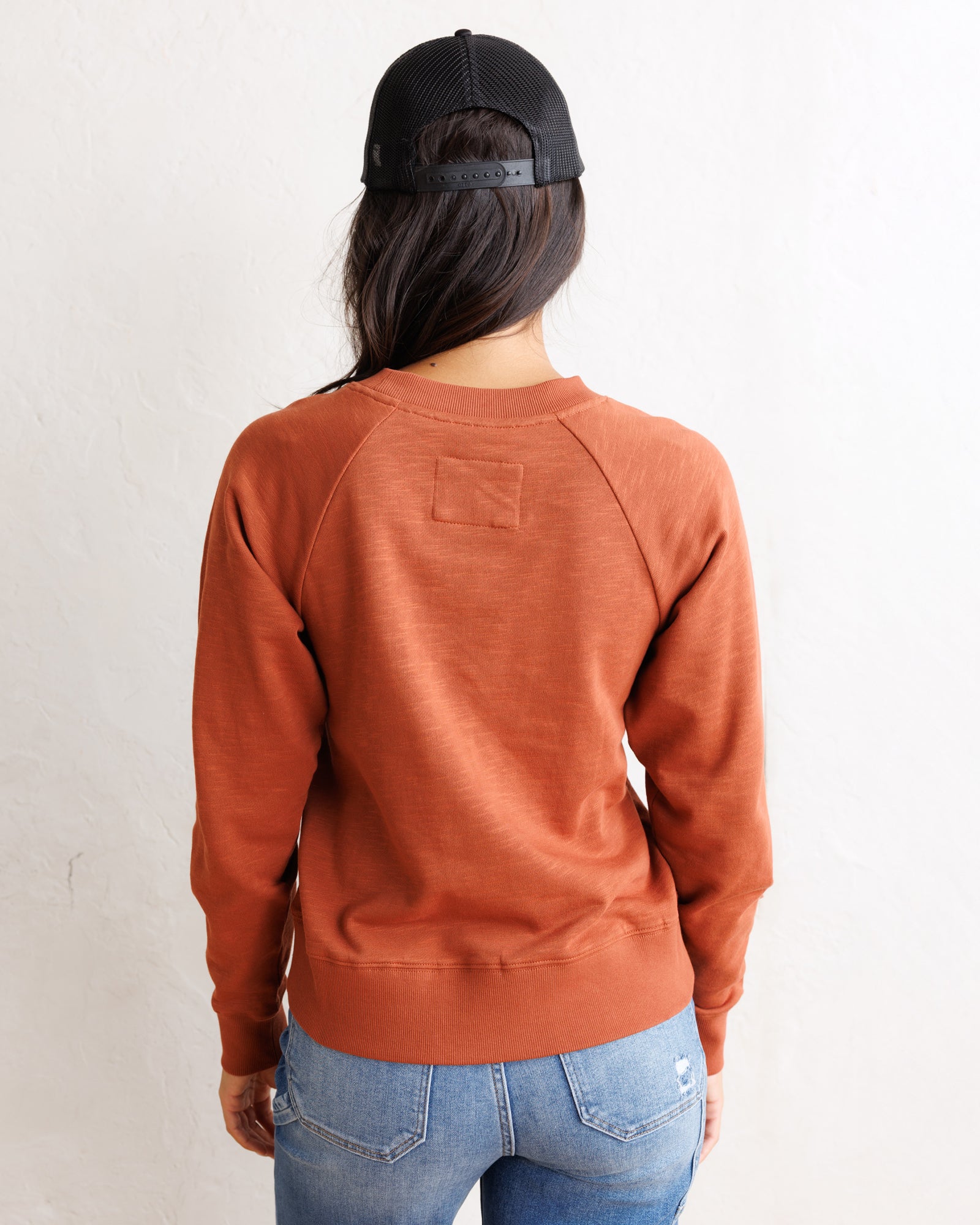 A woman viewed from behind wearing a rust-orange raglan crewneck sweatshirt, a black trucker hat, and blue jeans against a plain white wall.