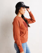 A woman wearing a rust-orange long-sleeve sweatshirt with a dark red heart patch on the chest, blue jeans, and a black baseball cap, posing against a textured off-white wall.