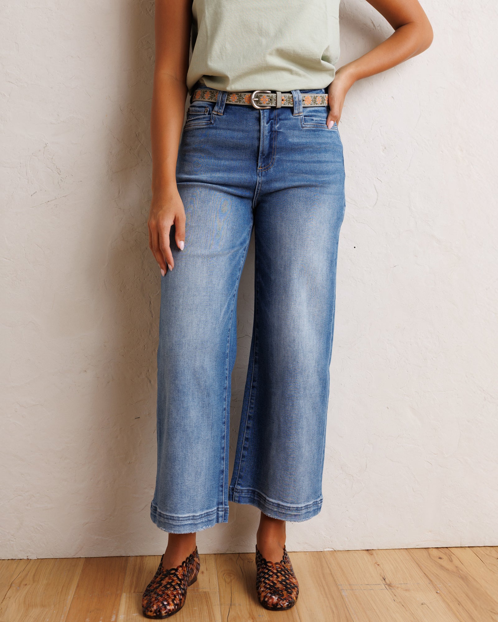 A model wearing a pair of wide-leg cropped blue denim jeans with a brown belt, paired with a light sage green top and leopard print flat shoes, standing against a textured off-white wall.