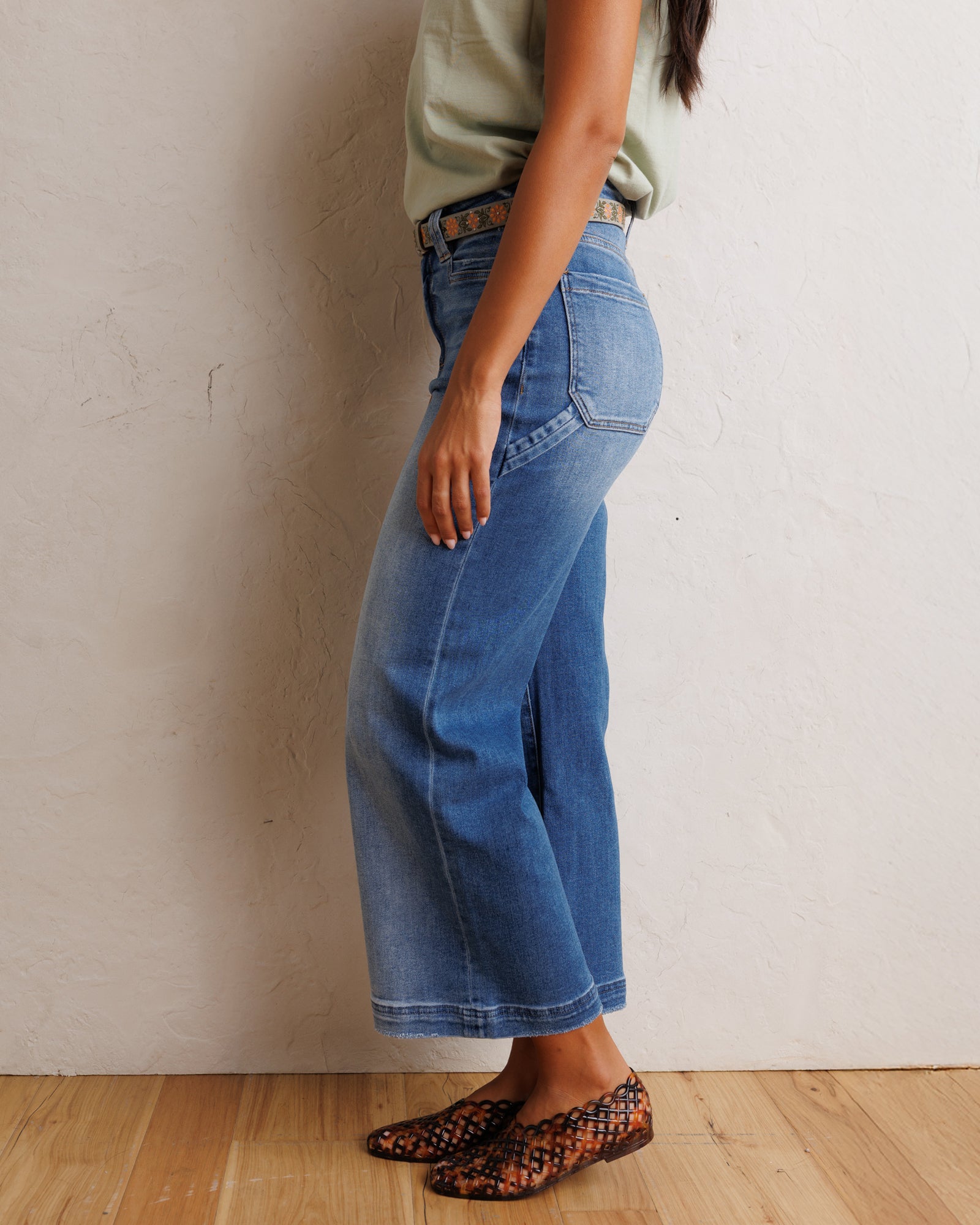 A person wearing blue wide-leg cropped jeans, a light sage green sleeveless top, and brown and black snakeskin-patterned flat shoes, standing against a textured off-white wall on a light wood floor.
