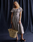 Woman in a checkered dress holding a woven bag against a dark background