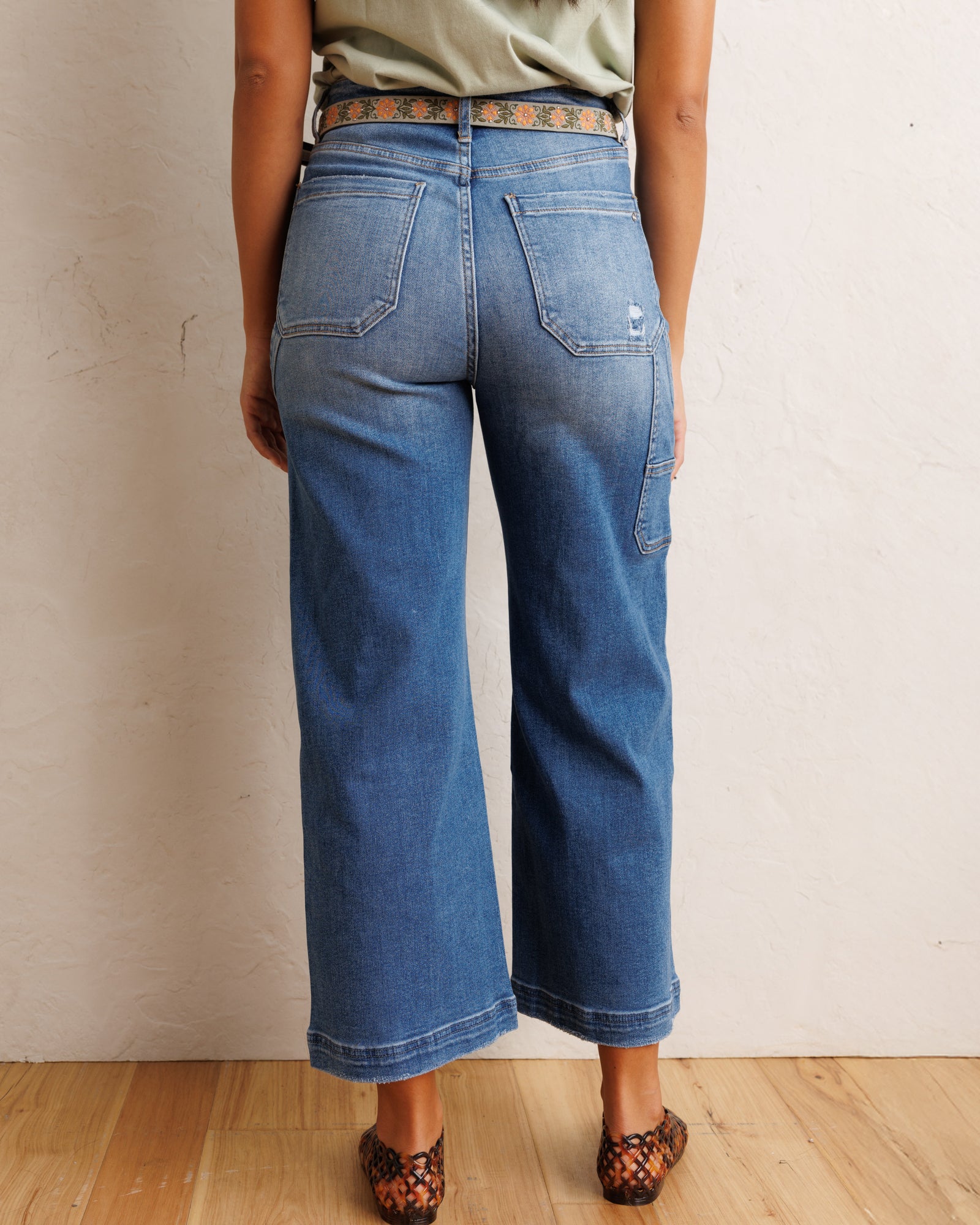 Rear view of a person wearing blue wide-leg cropped jeans with a patterned belt, olive top, and leopard-print shoes against a textured off-white wall and wood floor.