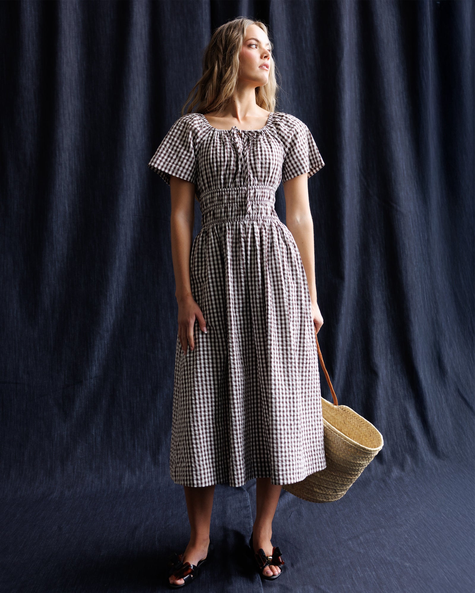 Woman in a checkered dress holding a woven basket against a dark blue curtain.