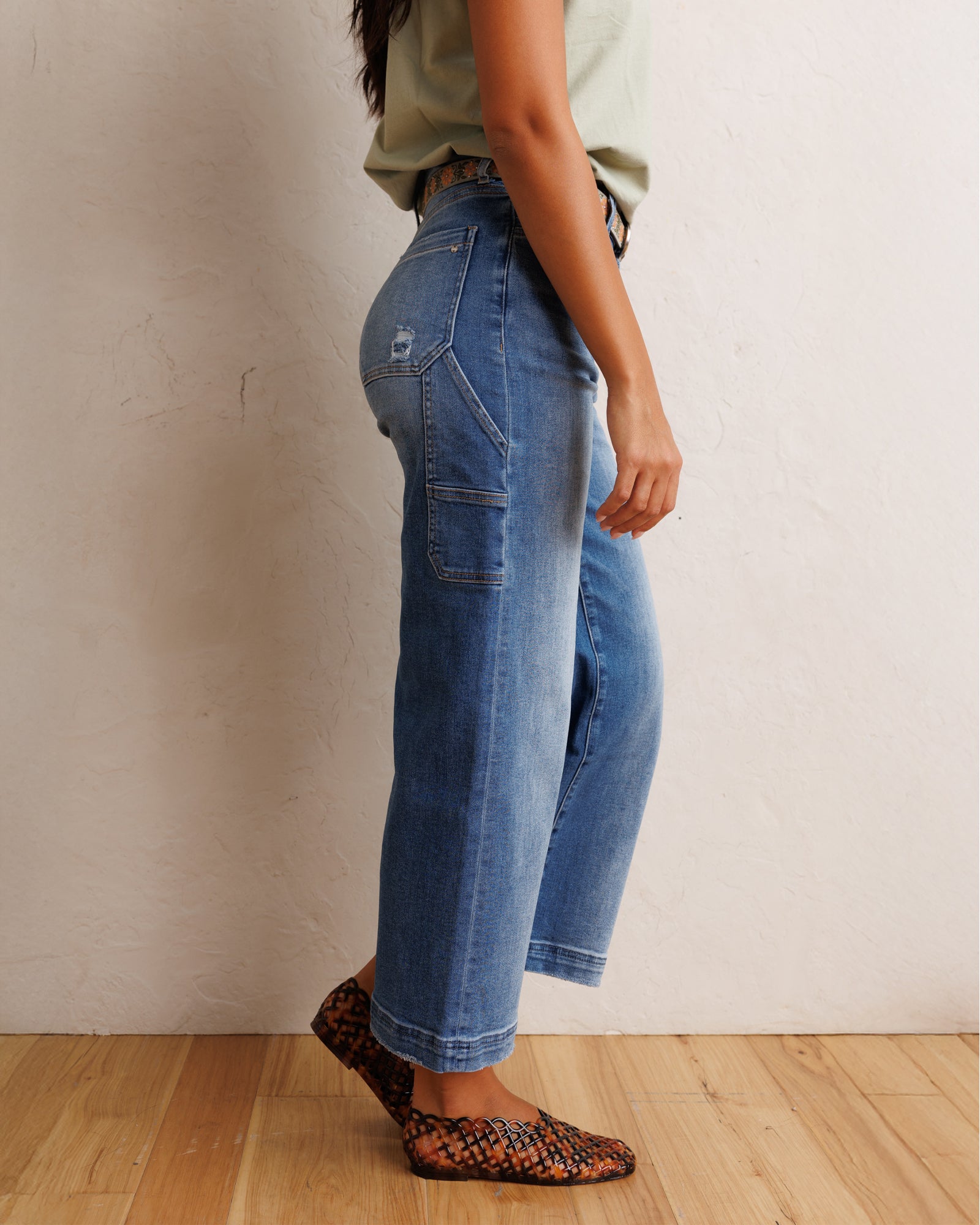 A person wearing blue wide-leg cropped jeans, a light sage green sleeveless top, and brown and black snakeskin-patterned flat shoes, standing against a textured off-white wall on a light wood floor.