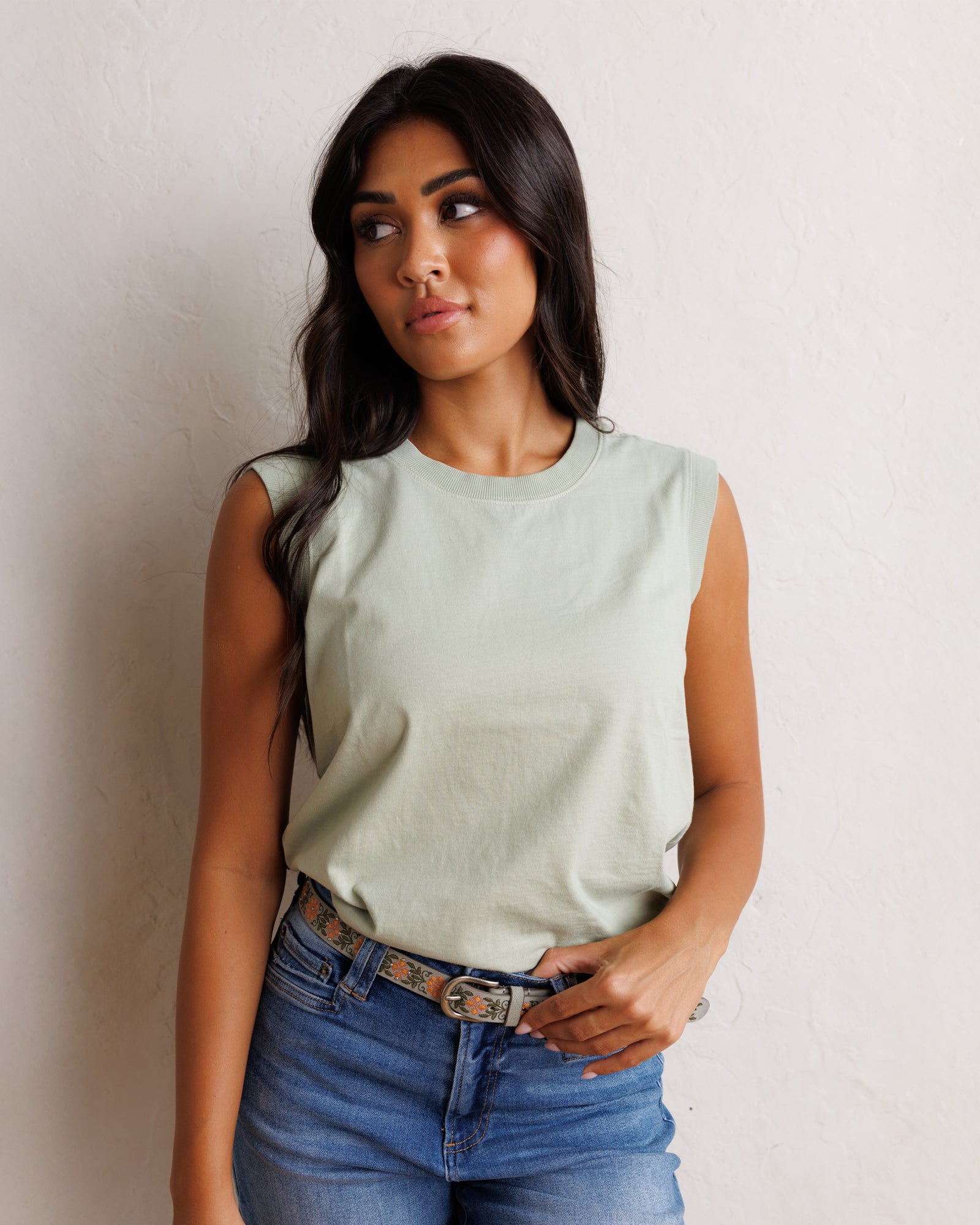 A woman wearing a sage green sleeveless crop top and blue jeans, posing against a textured off-white wall.