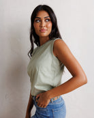 A woman wearing a sage green sleeveless crop top and blue jeans, posing against a textured off-white wall.