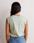 A woman viewed from behind wearing a light sage green sleeveless tank top and blue jeans, standing against a textured off-white wall.
