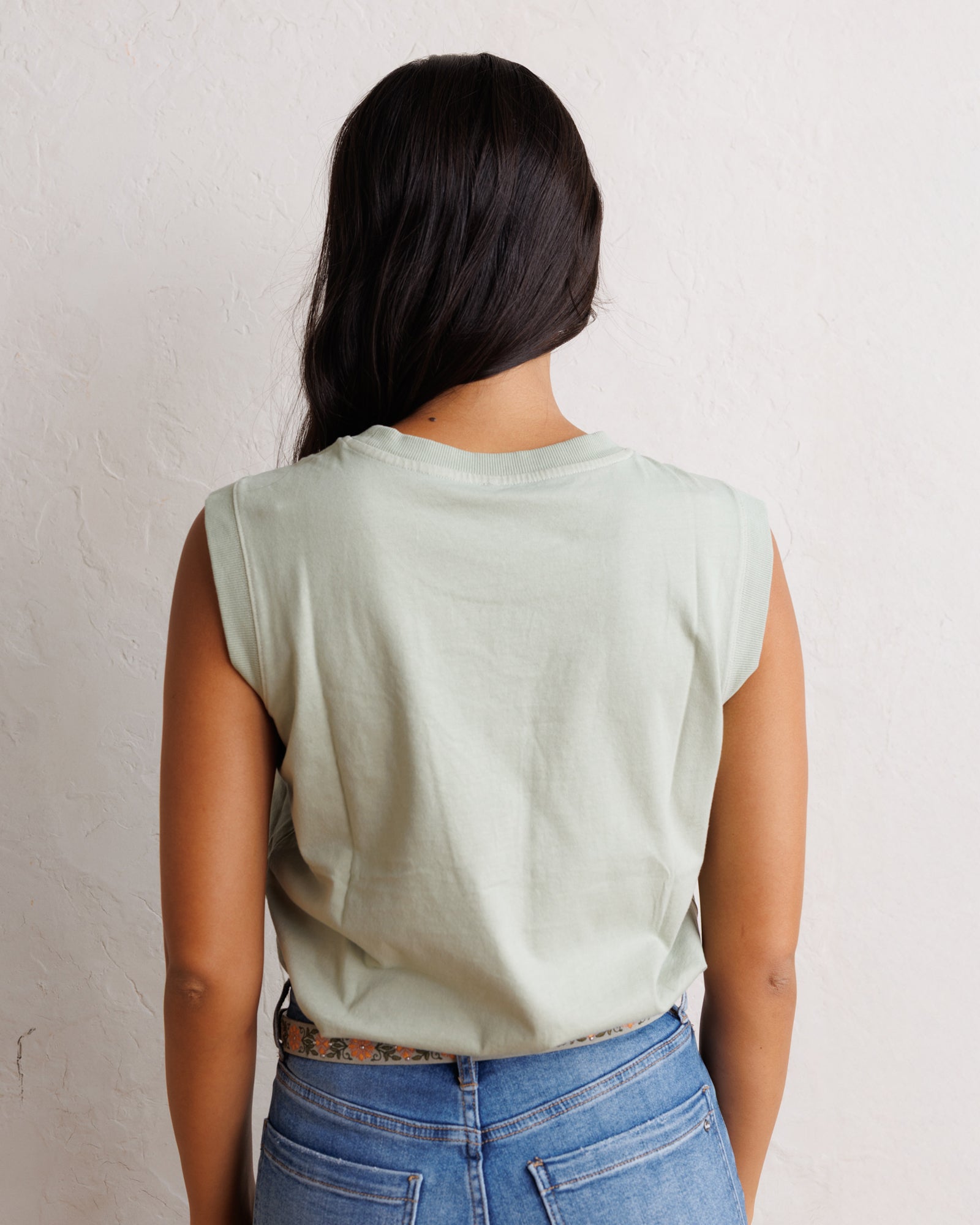 A woman viewed from behind wearing a light sage green sleeveless tank top and blue jeans, standing against a textured off-white wall.