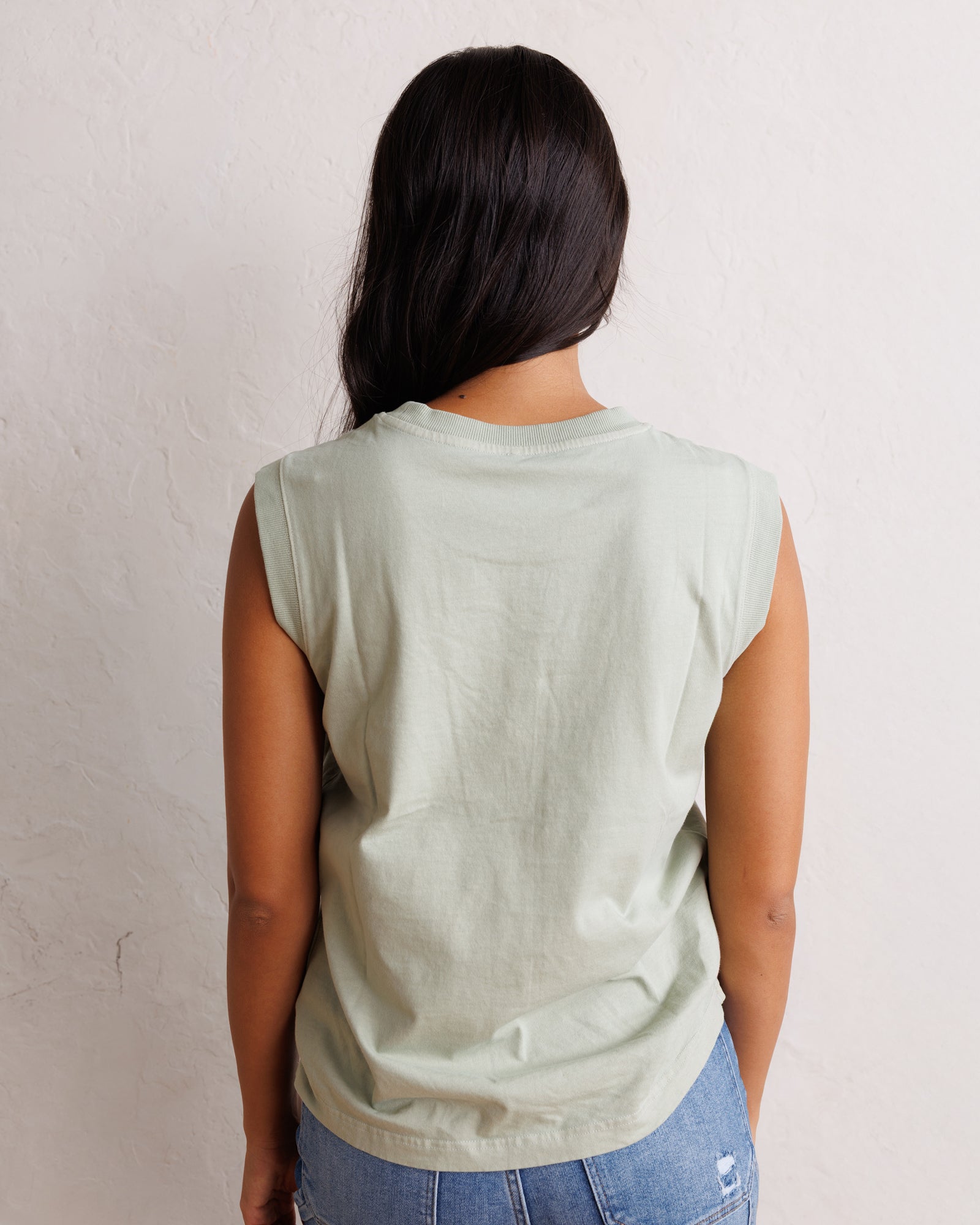 A woman viewed from behind wearing a light sage green sleeveless t-shirt and blue jeans, standing against a textured off-white wall.