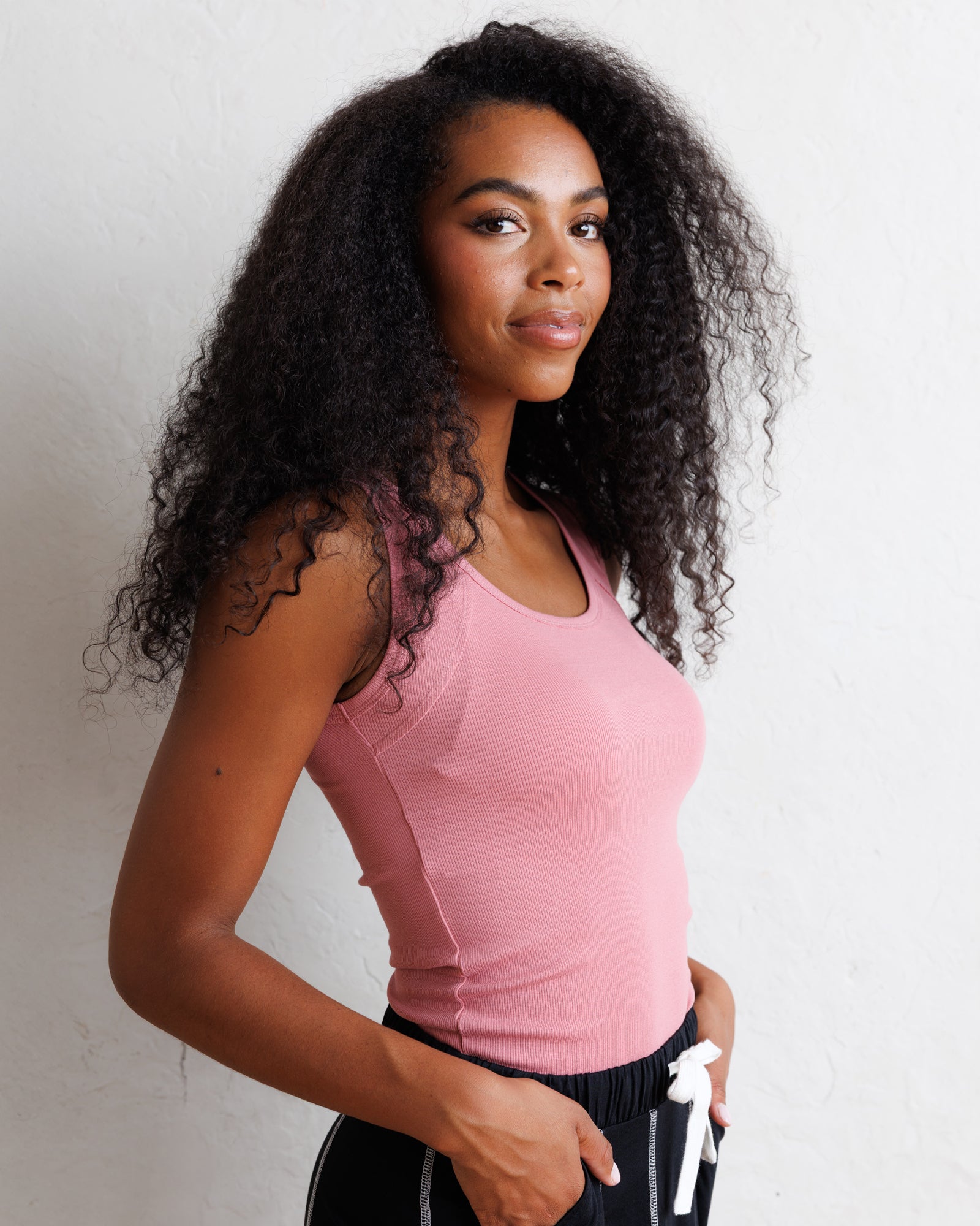 A woman with long, curly black hair wearing a fitted light pink athletic tank top and black athletic pants with a white drawstring, posing against a white textured wall.