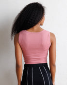 A woman shown from behind wearing a fitted sleeveless pink top and black pants with thin white vertical stripes, standing against a plain off-white wall.