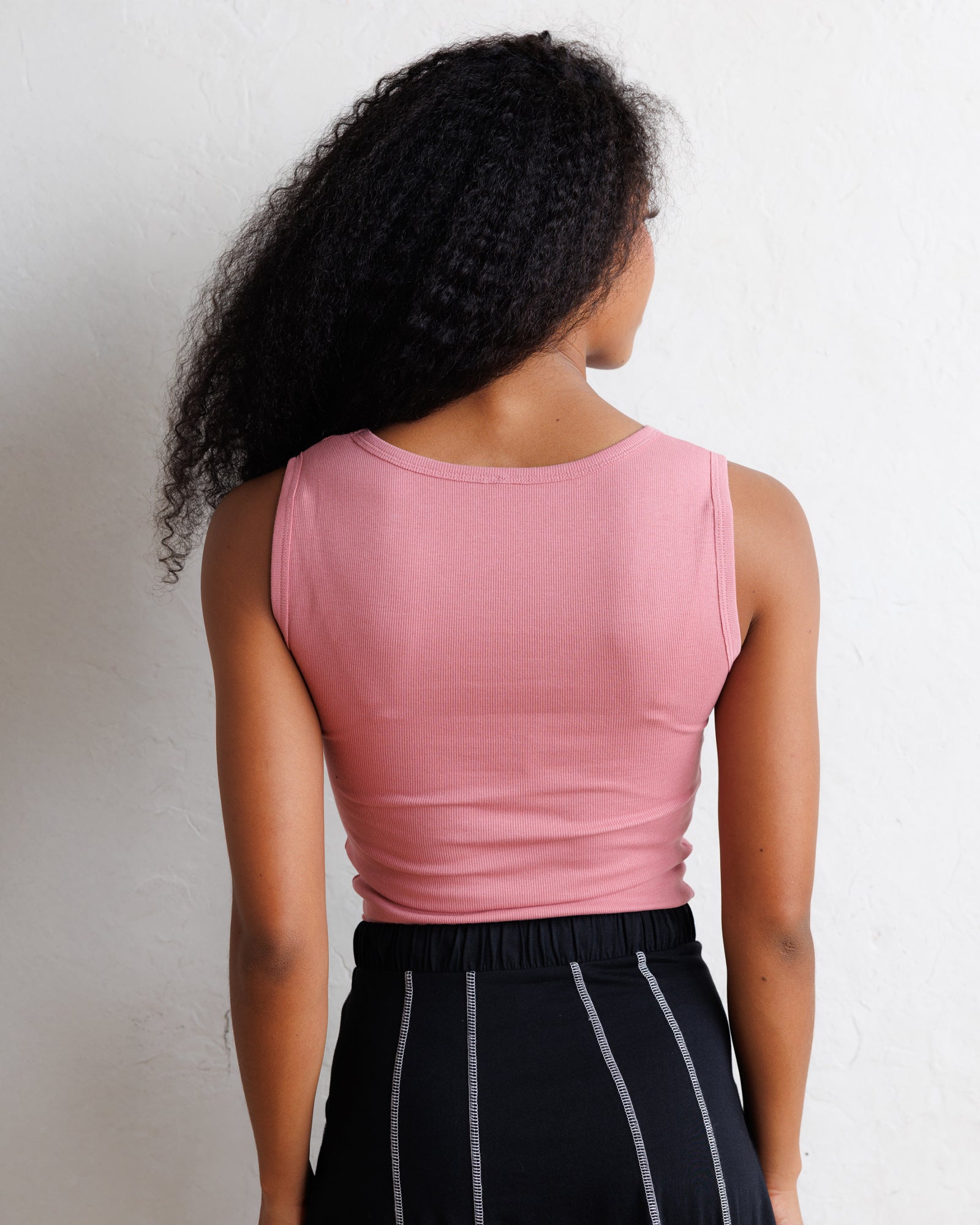 A woman shown from behind wearing a fitted sleeveless pink top and black pants with thin white vertical stripes, standing against a plain off-white wall.