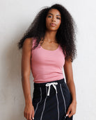 A woman with long, curly black hair wearing a pink ribbed tank top and dark navy pants with white vertical stripes and a white drawstring, posing against a textured off-white wall.