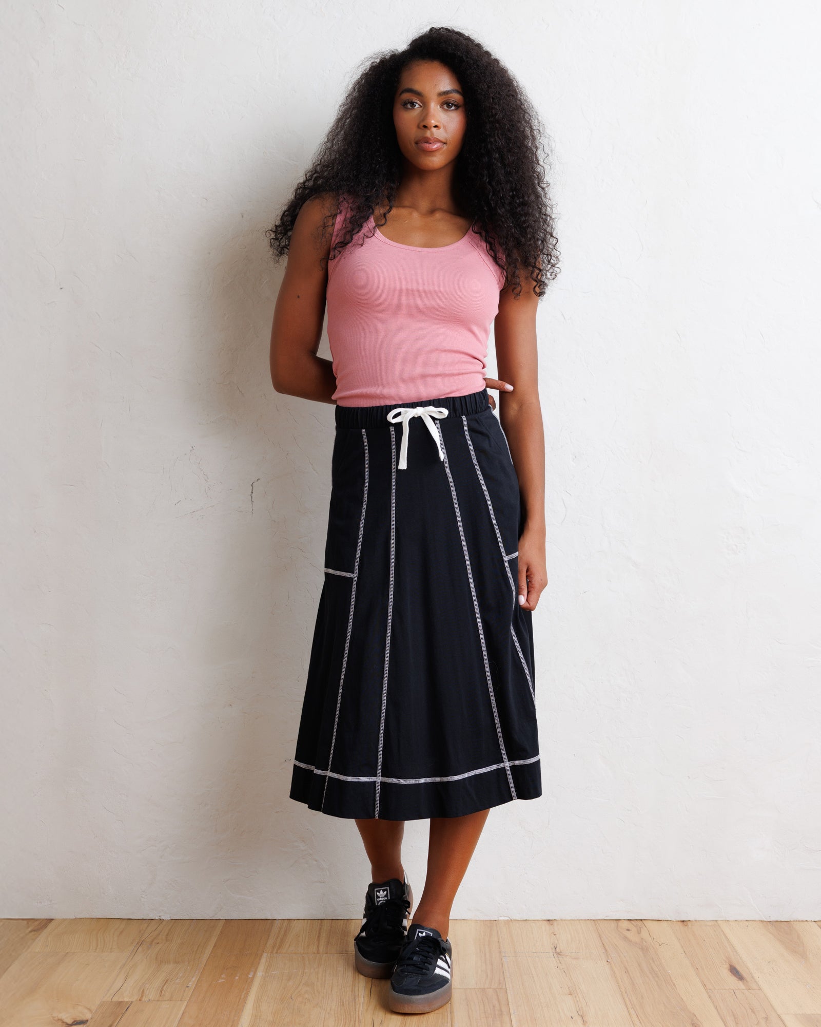 A woman stands against a light textured wall on a wood floor, wearing a pink sleeveless tank top and a black midi skirt with white vertical stripe lines and a white drawstring. She is wearing black sneakers.
