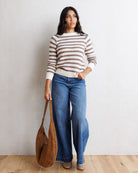 A woman stands against a white wall on a light wood floor, wearing a beige and brown horizontally striped knit sweater, blue wide-leg jeans, and holding a brown studded shoulder bag.