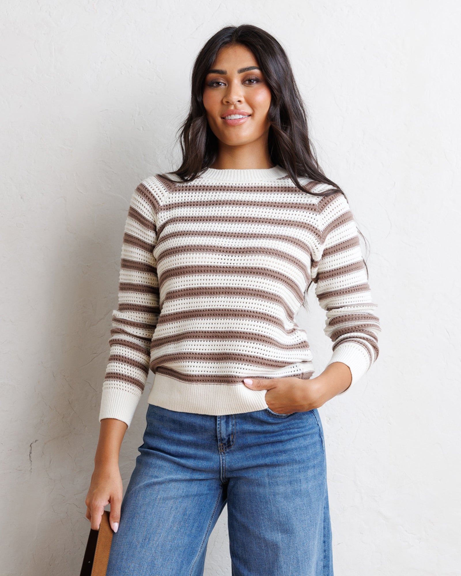 A woman wearing a brown and cream striped knit sweater with blue jeans, standing against a textured off-white wall.