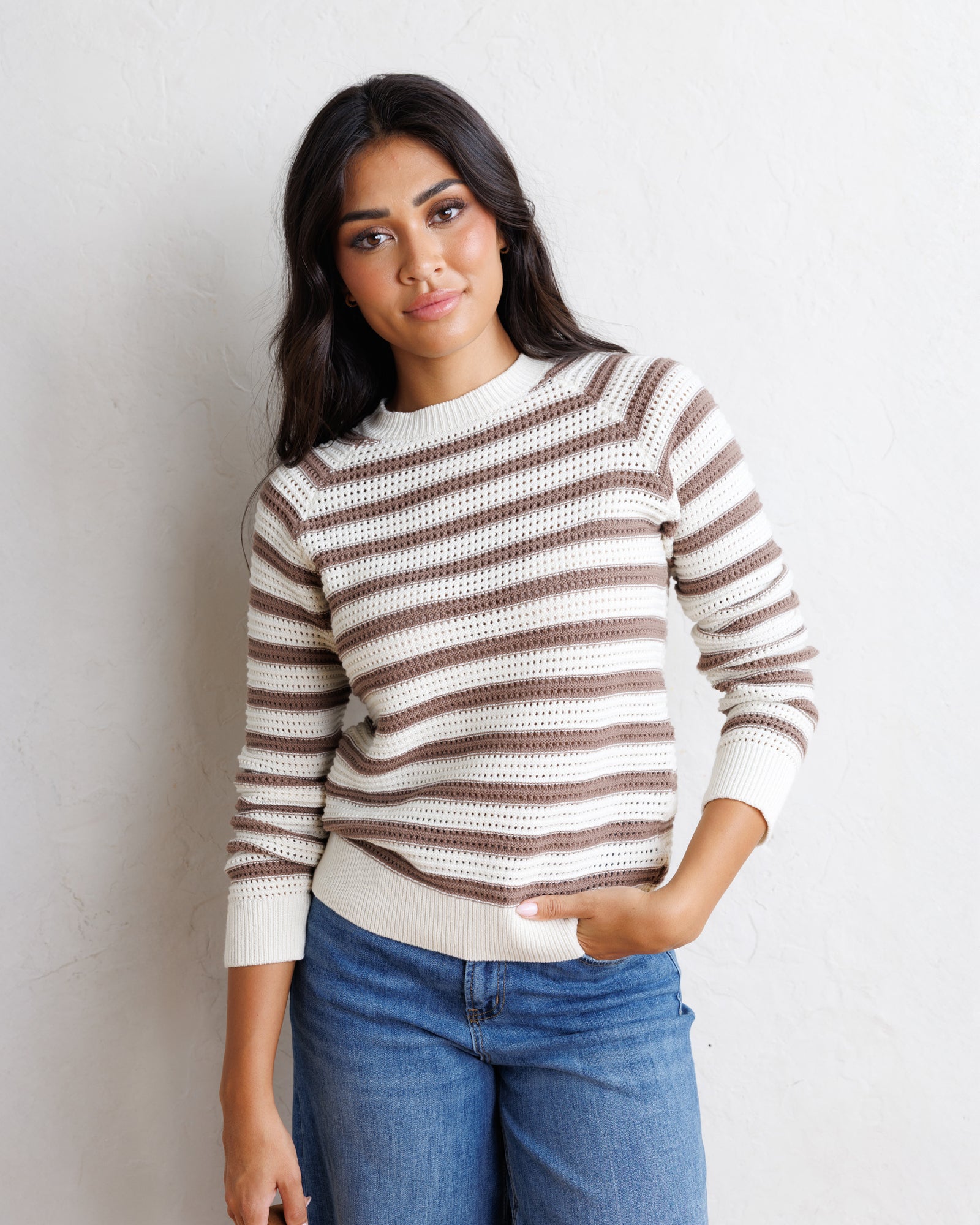 A woman wearing a brown and cream striped knit sweater with blue jeans, standing against a textured off-white wall.