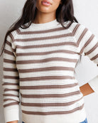 A woman wearing a knit crewneck sweater with horizontal brown and cream stripes, shown from the chest up against a light neutral wall.