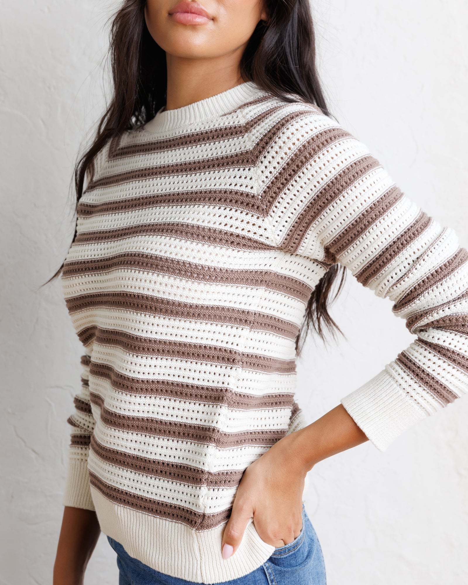 A woman wearing a cream and brown striped knit sweater with a textured openwork pattern, paired with blue jeans, against a light textured wall.