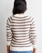 A woman viewed from behind wearing a cream and brown horizontally striped knit sweater with a textured openwork pattern, paired with blue jeans against a plain white wall.
