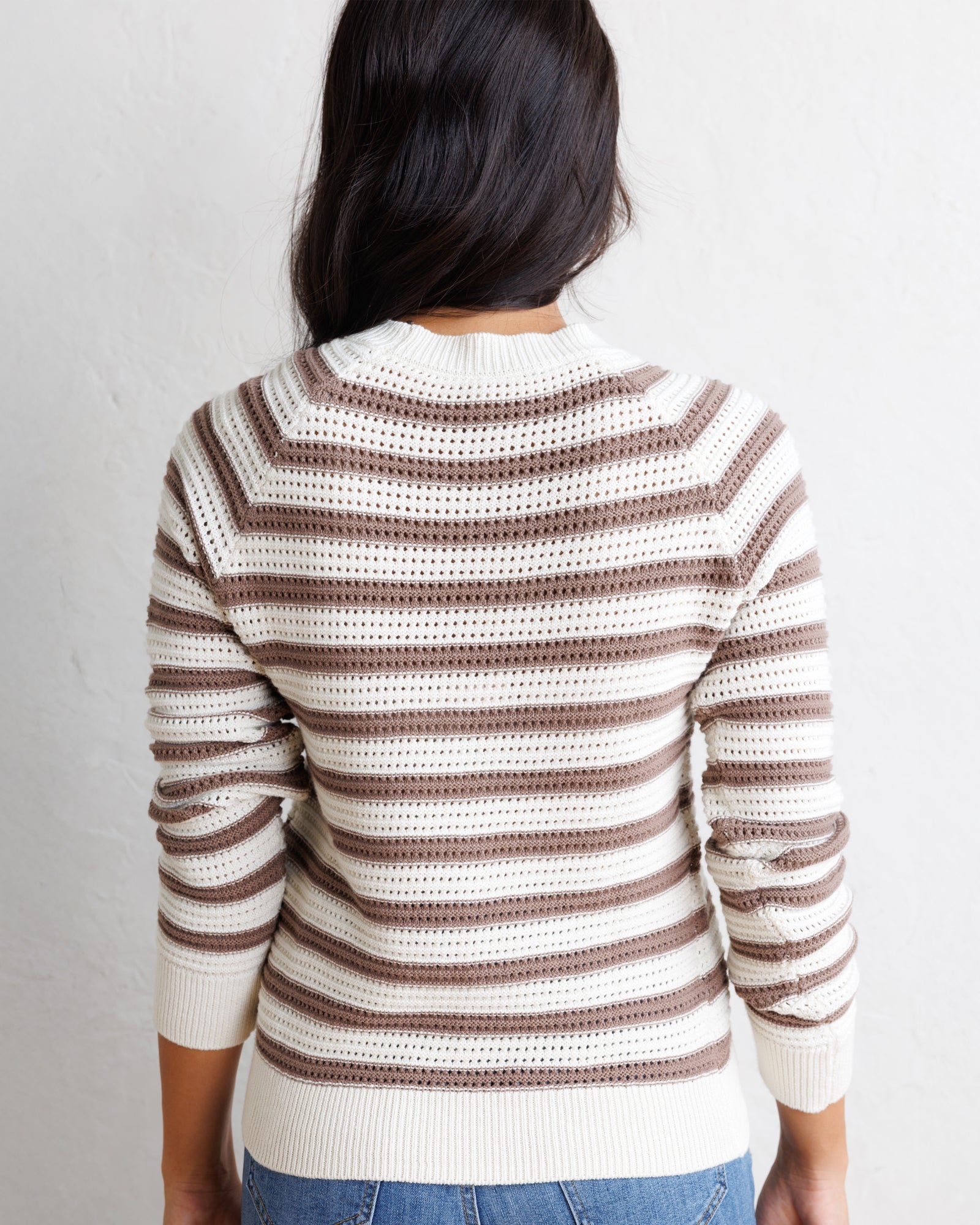 A woman viewed from behind wearing a cream and brown horizontally striped knit sweater with a textured openwork pattern, paired with blue jeans against a plain white wall.