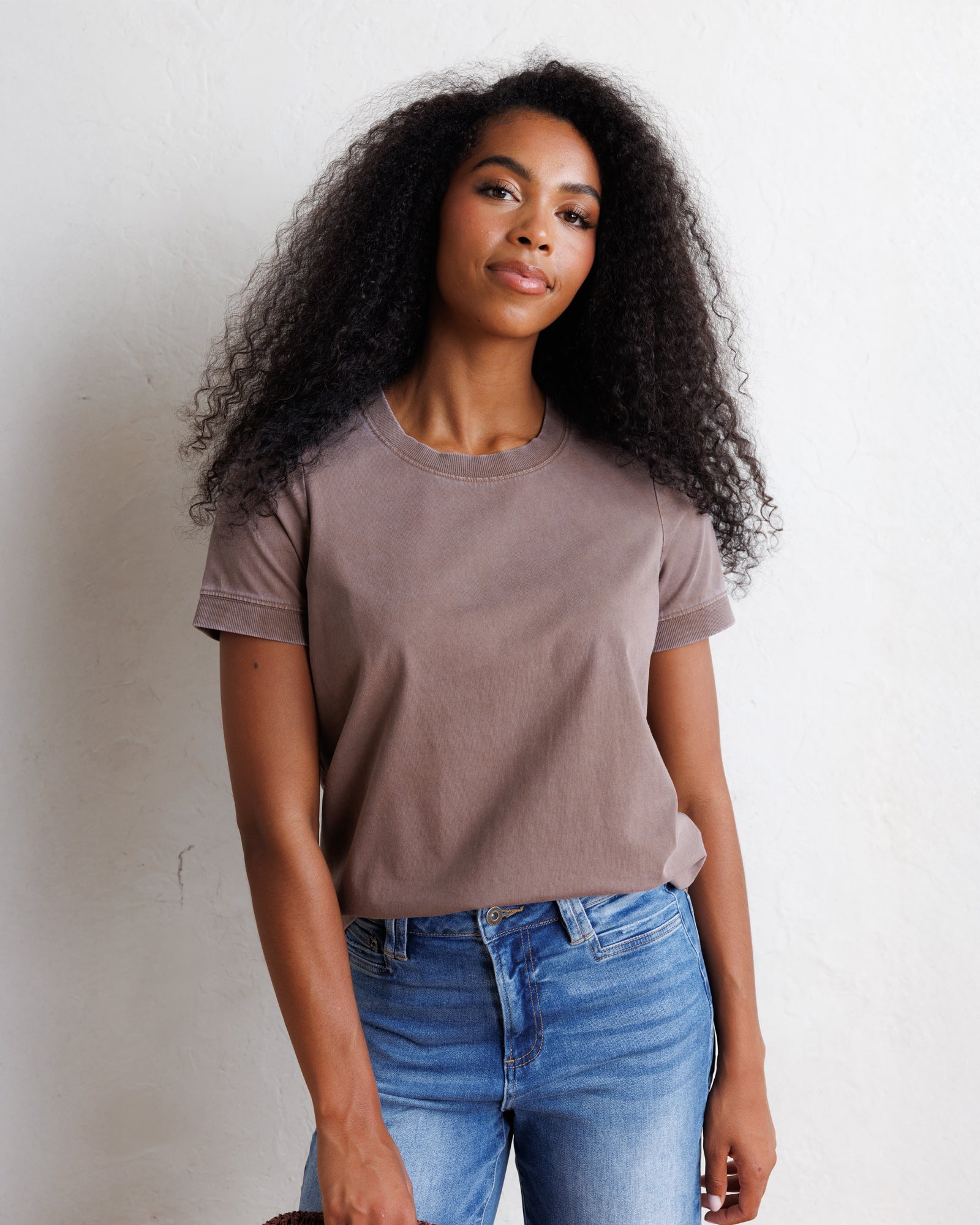 A woman with long, curly black hair wearing a muted taupe-brown short-sleeve crewneck t-shirt and blue jeans, standing against a textured off-white wall.