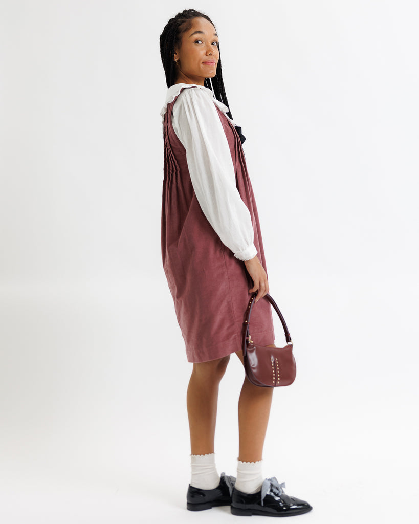 Girl wearing a burgundy pinafore dress with a white shirt underneath, holding a brown handbag on a white background.