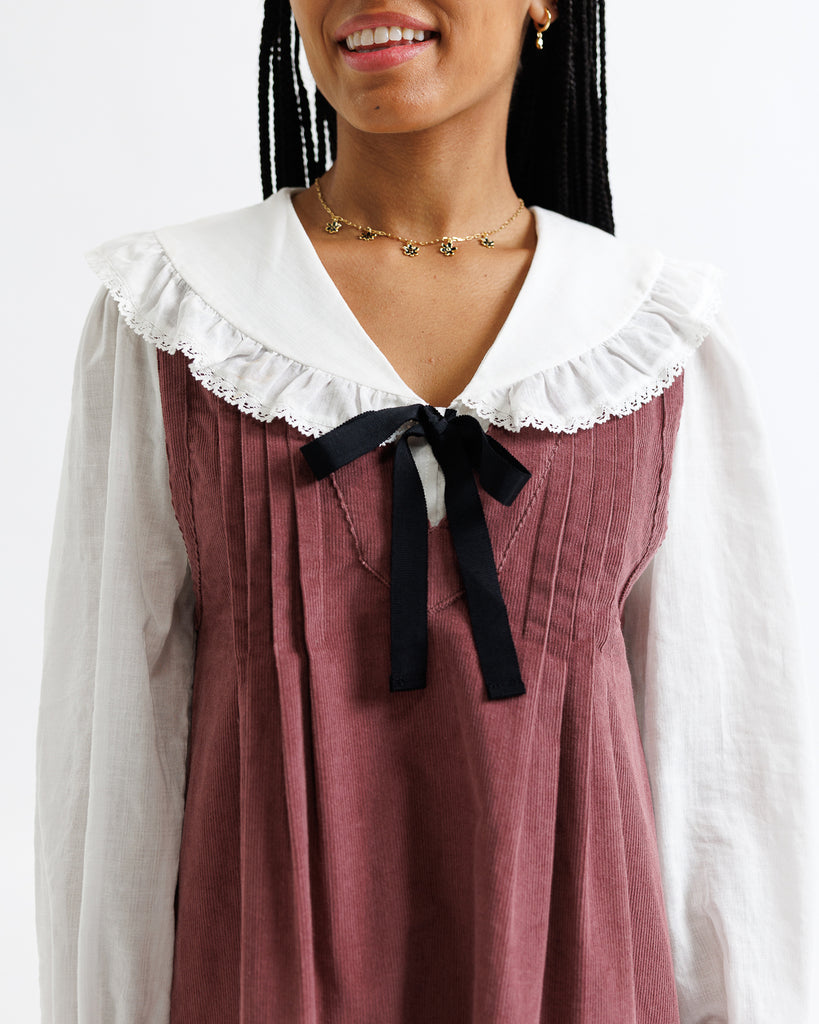 Person wearing a maroon dress with a white collar and black ribbon, standing against a plain background.